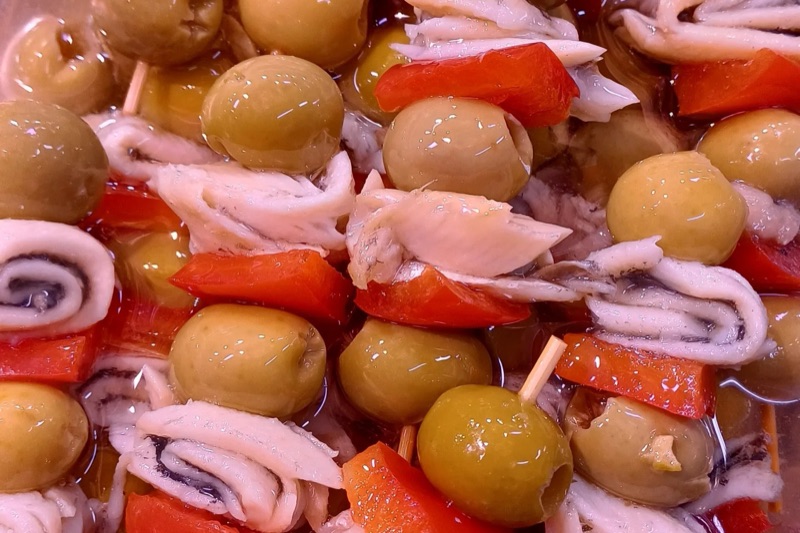 Bandeja densa de pinchos del puesto: aceitunas, anchoas enrolladas y pimiento rojo en vinagre.