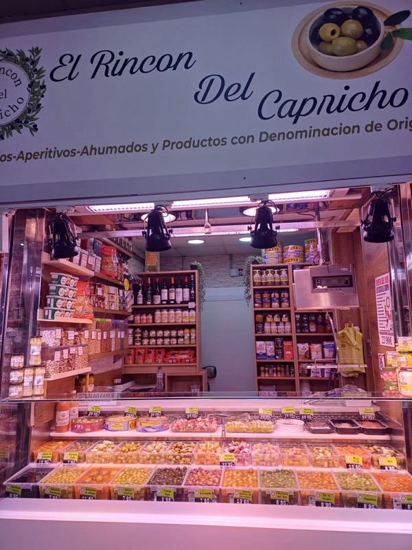 Vista frontal del puesto: vitrina con bandejas de aceitunas, gildas y encurtidos, y estantería de vinos al fondo bajo el cartel del puesto.