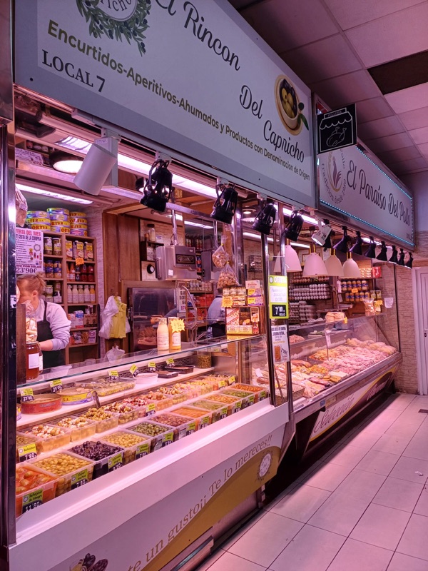 Vista del puesto desde el pasillo del Mercado de San Isidro, con la vitrina larga de encurtidos del Rincón del Capricho y el puesto vecino al fondo.